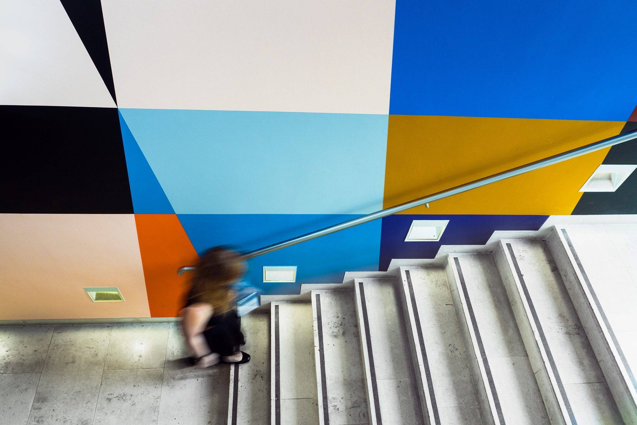 View looking down a stairwell decorated with a colourful geometric mural as a woman dressed in black climbs the stairs.