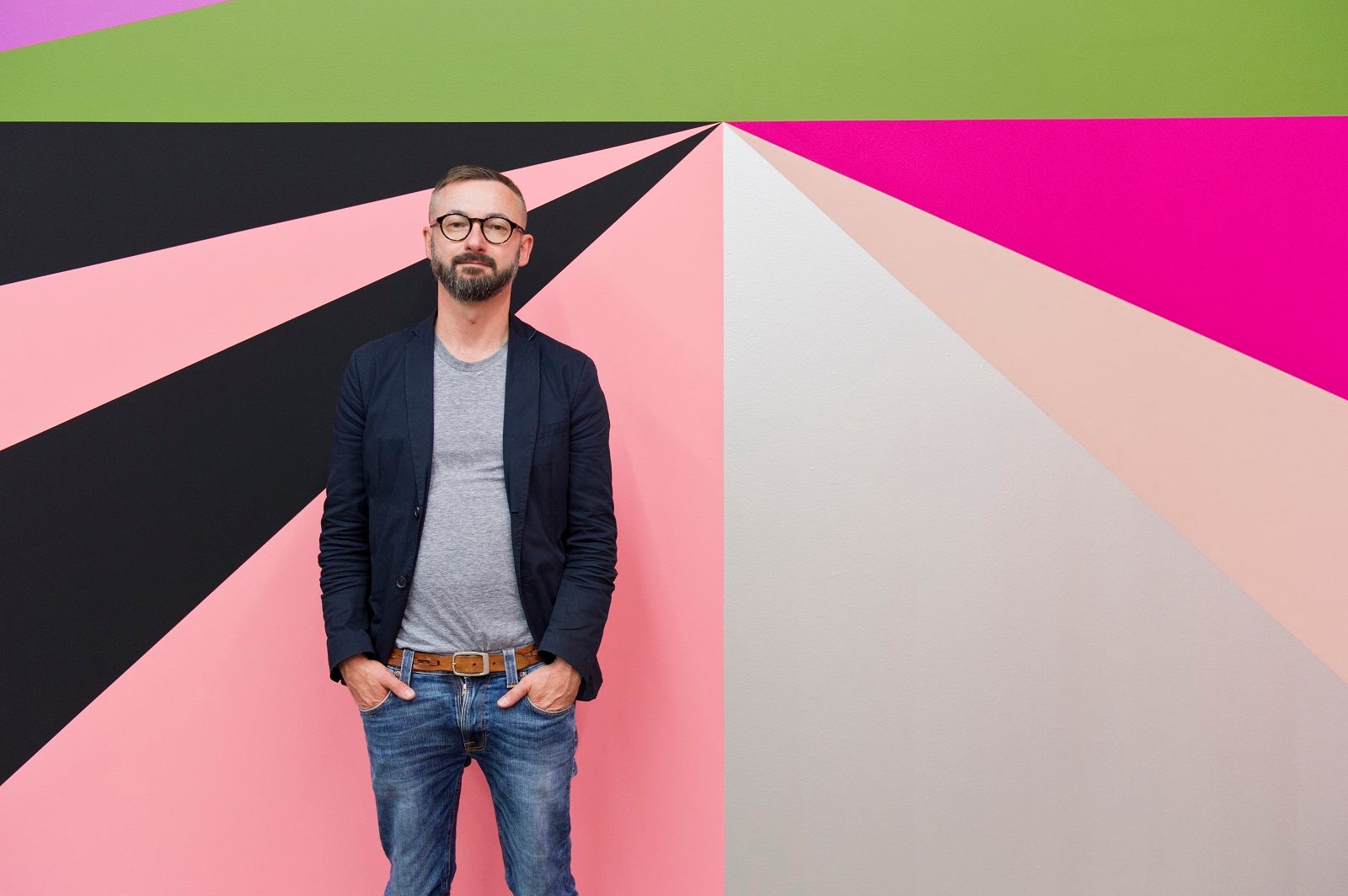 A man wearing a grey t-shirt, black jacket and blue jeans stands in front a of a mural of brightly coloured geometric shapes.