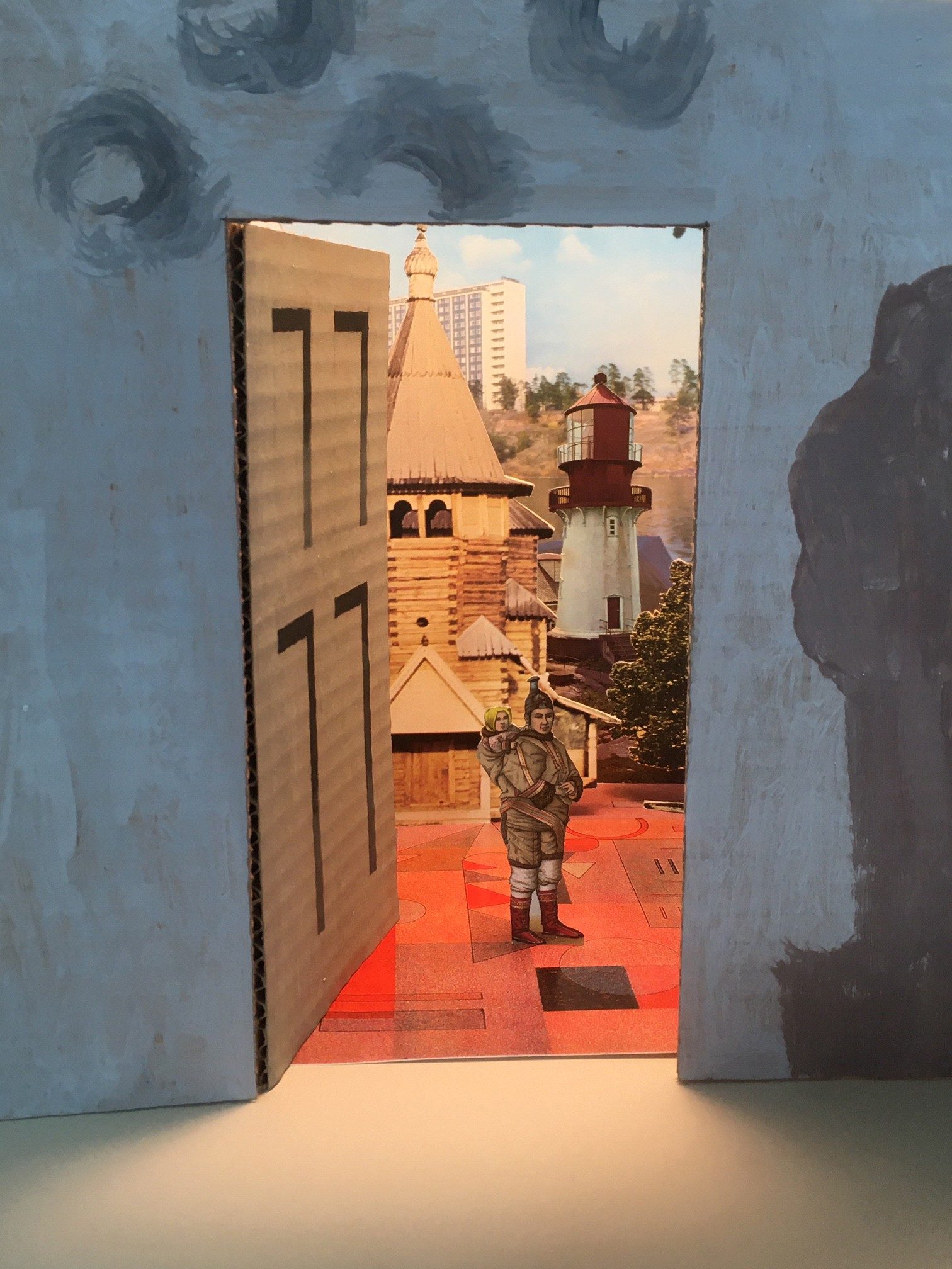 A piece of cardboard decorated to look like a wall with a door opens onto a surreal collage landscape showing a woman carrying a child with a variety of buildings in the background including a tower block and a lighthouse