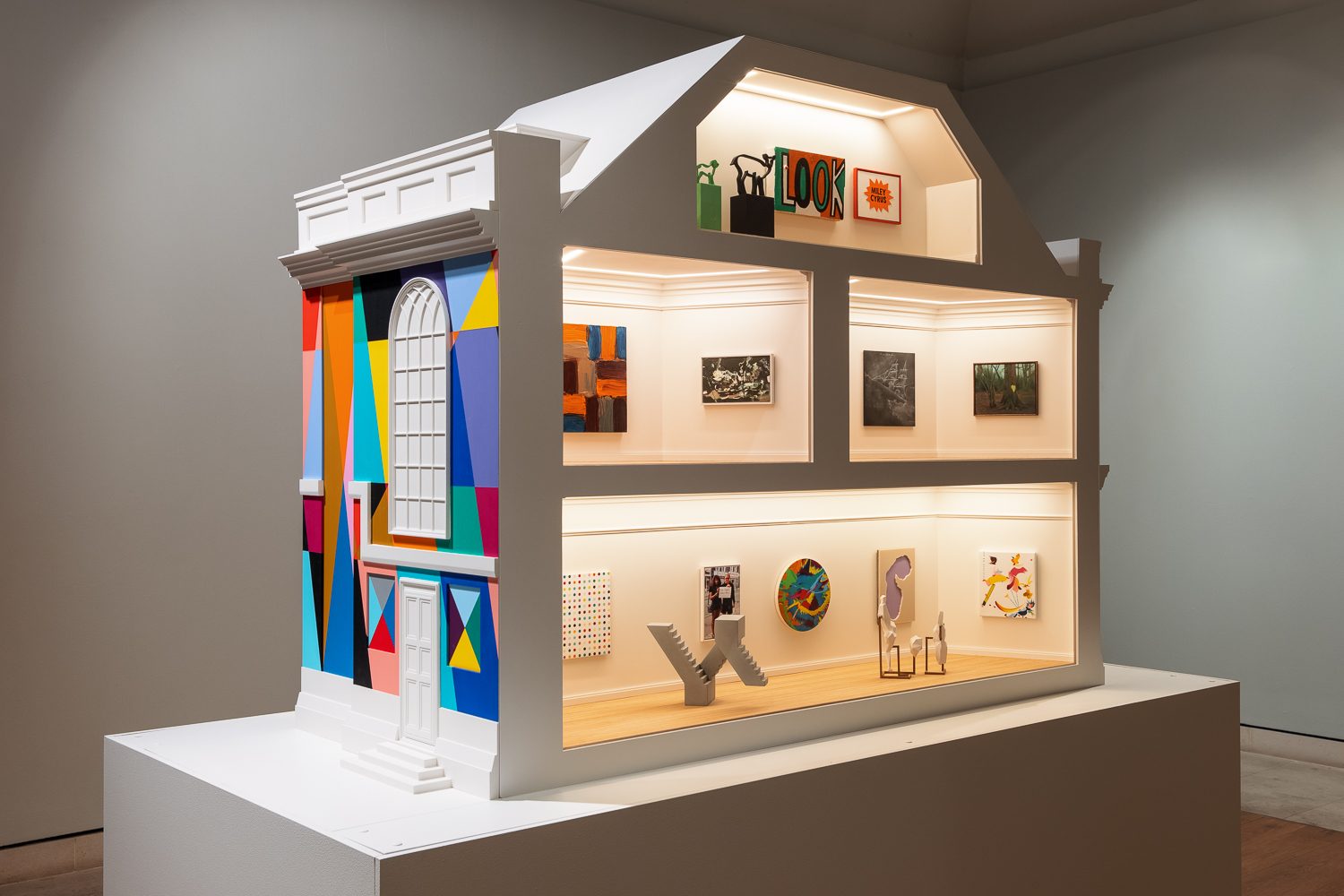 A large dollhouse sized art gallery, designed to imitate the exterior of an 18th century townhouse. The left hand side is painted with a brightly coloured geometric mural. The right side shows a cross section of four rooms (one in the attic, two on the first floor, and one on the ground floor), each filled with works of modern art.